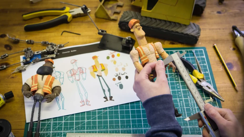 Hands measuring a stop-motion character puppet on a crafting mat, surrounded by tools and character sketches, illustrating the detailed handmade process of stop-motion animation.